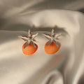 Sunset Shell statement earrings featuring textured coral shell drops with white floral tops and pearl centers displayed on ivory satin fabric.