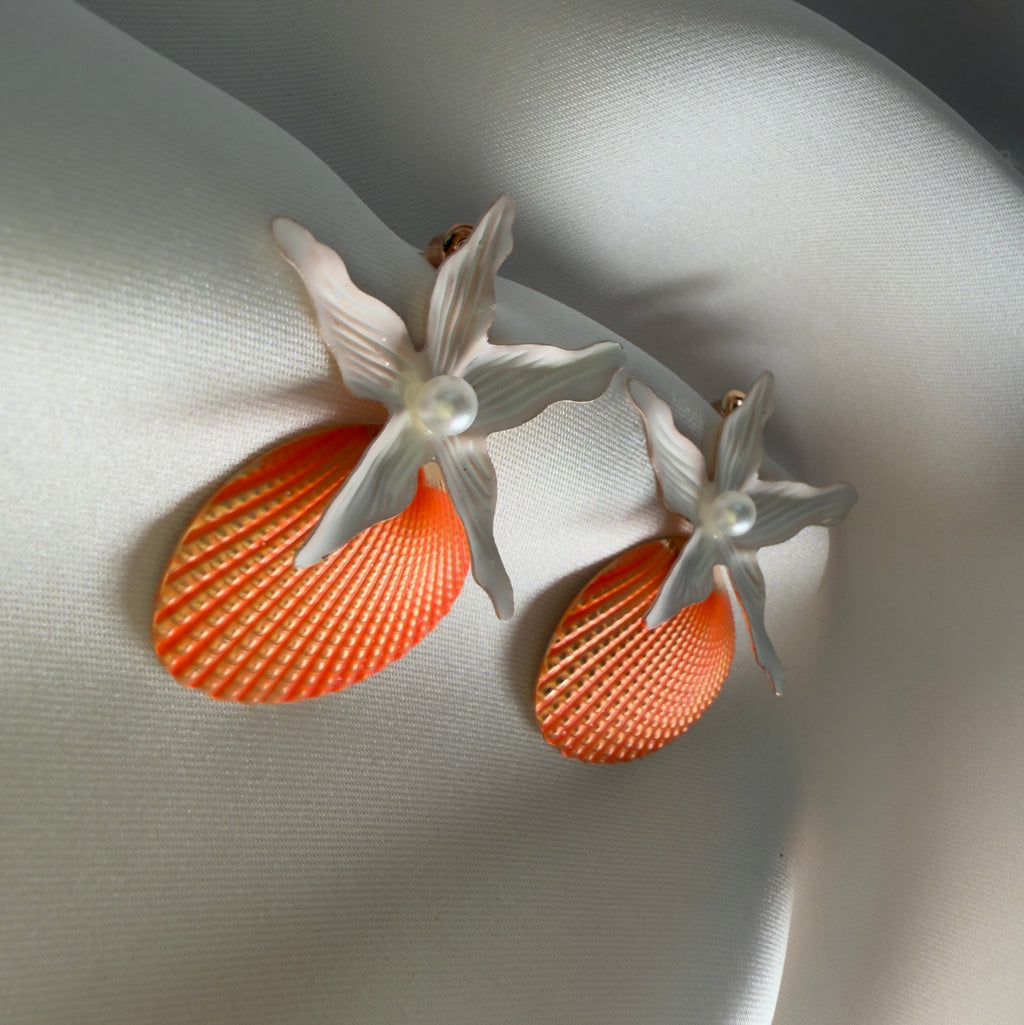 Close-up of coral blossom statement earrings with textured orange shell design, white flower detail, and pearl center on soft ivory satin.